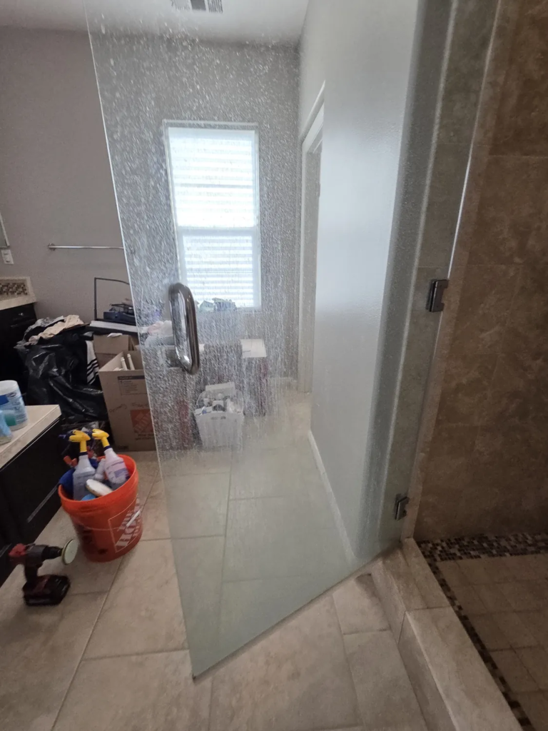 Frameless shower door before service, clouded with white hard-water streaks so the window and room behind it are blurred