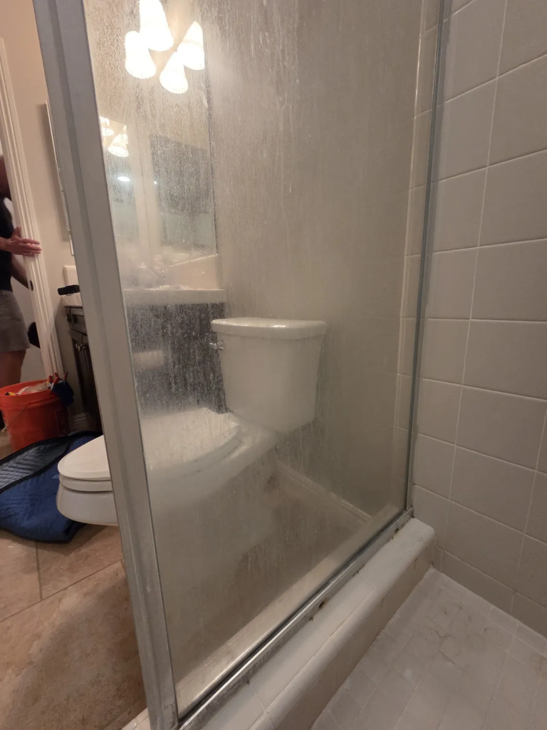 Fixed shower panel before restoration, with dense white water spots obscuring the toilet behind it