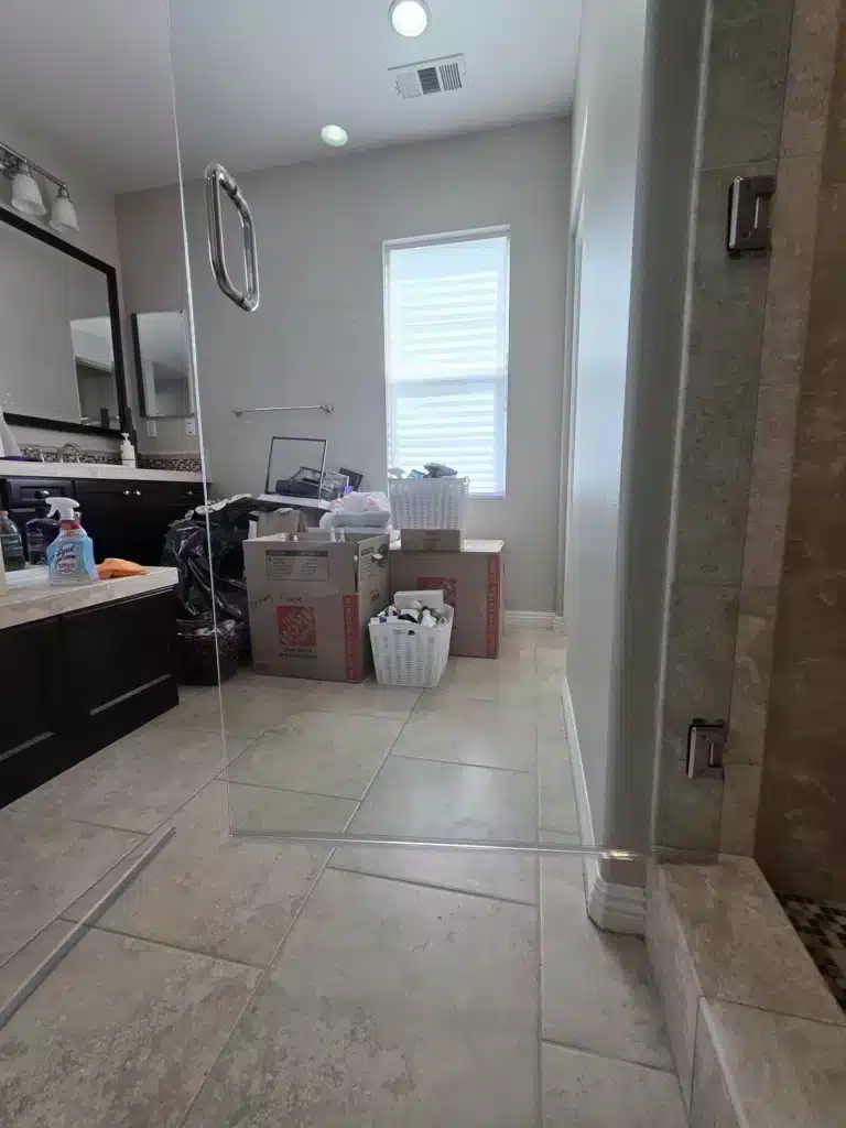 The same frameless shower door after restoration, clear, with the bathroom vanity and window visible behind it