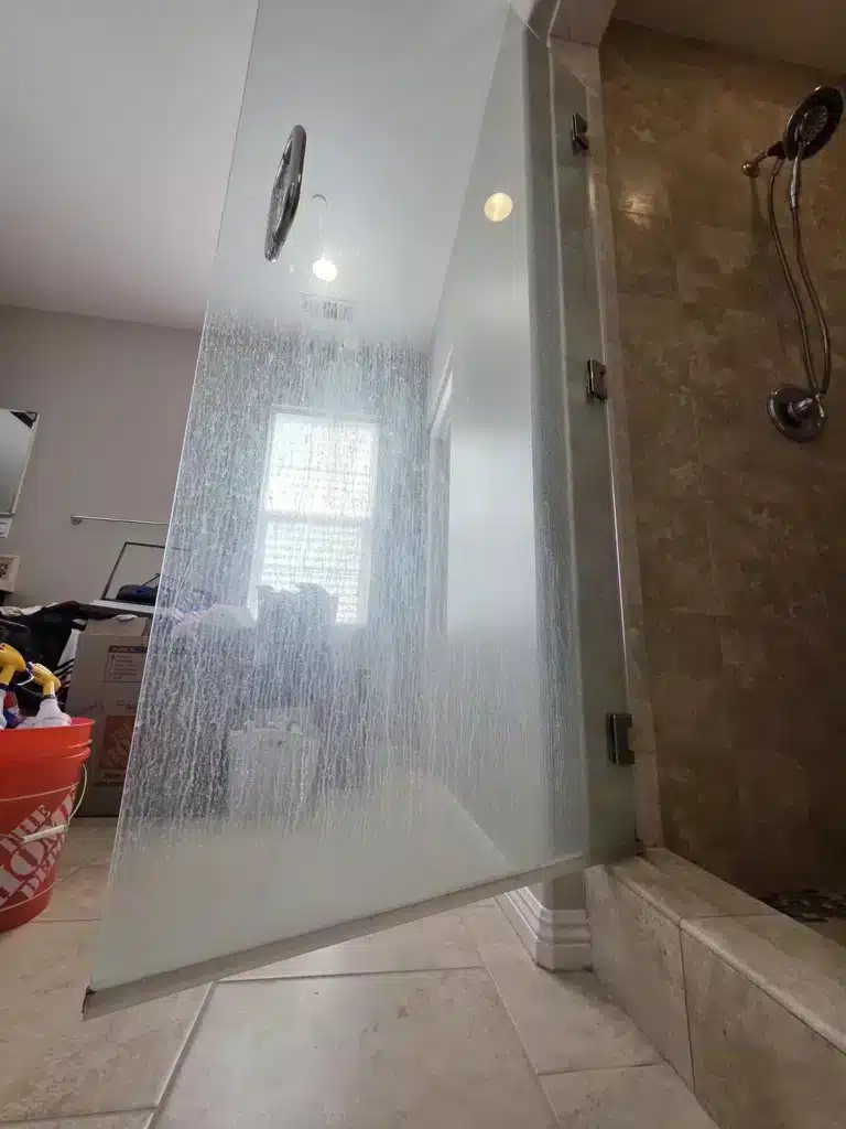 Frameless shower door before restoration, covered in white hard-water streaks so the room behind it is barely visible